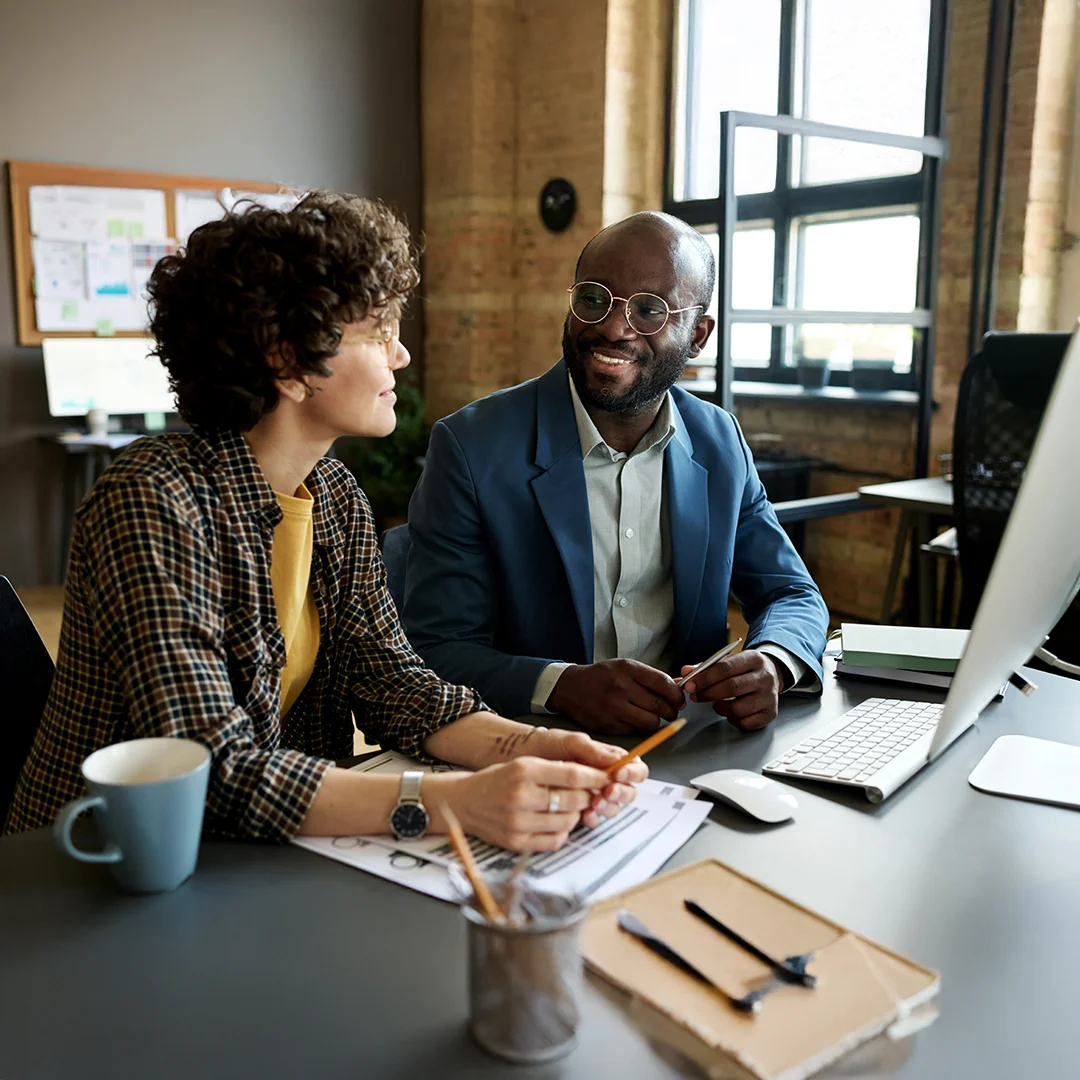 Two coworkers having a discussion at a desk with papers and a computer.