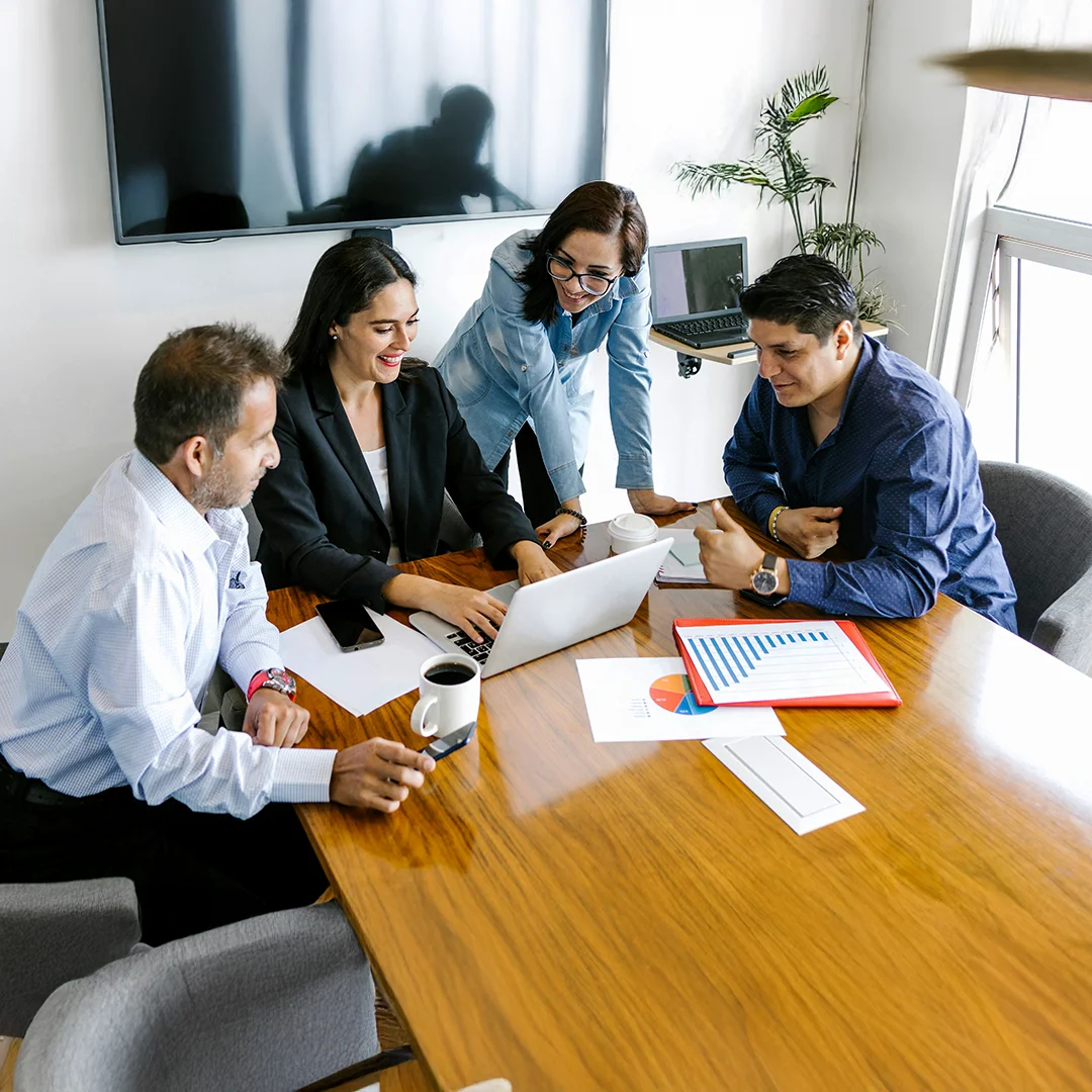 Four coworkers gathered at a table discussing data on a laptop and printed charts.