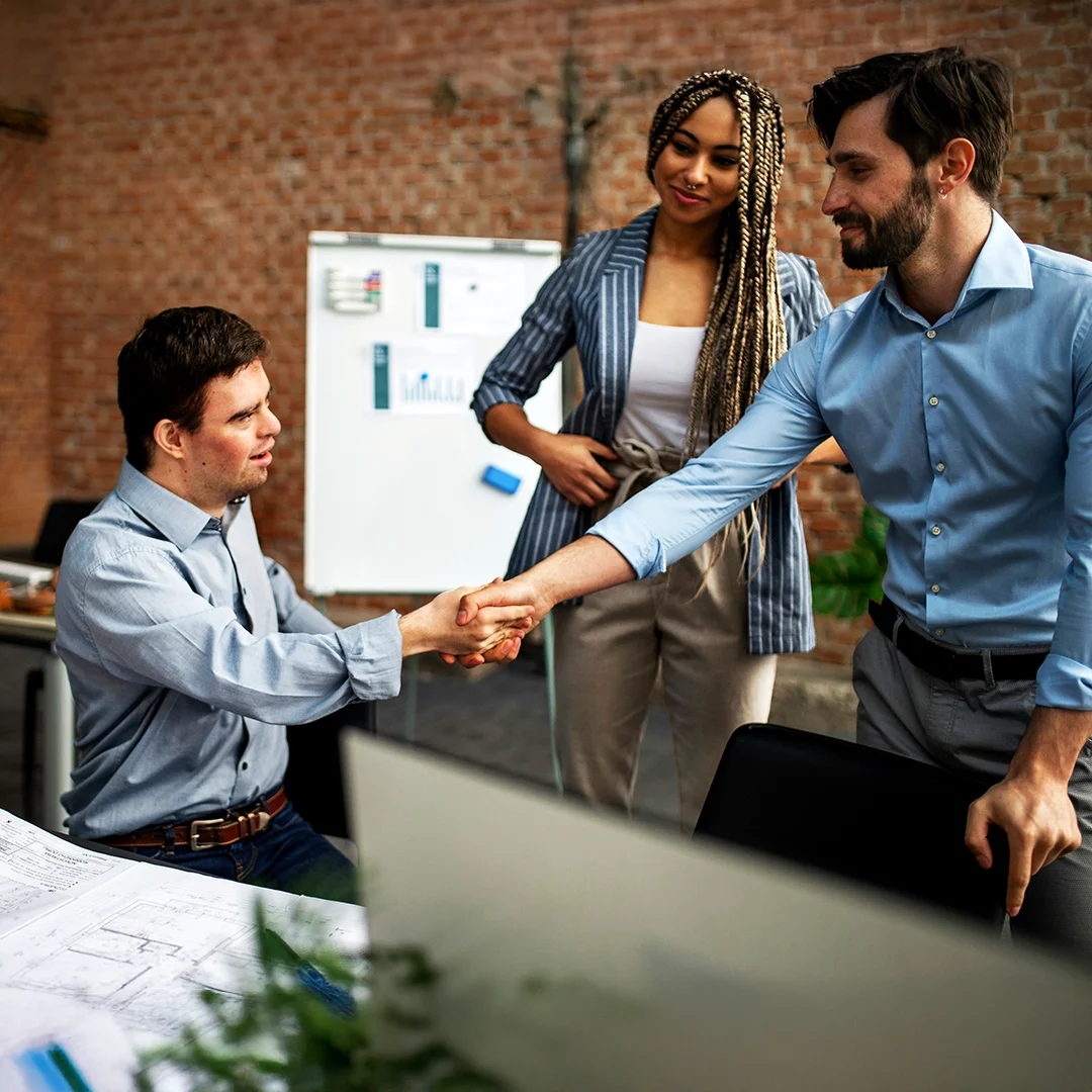 Three coworkers in an office as two of them shake hands near architectural plans.