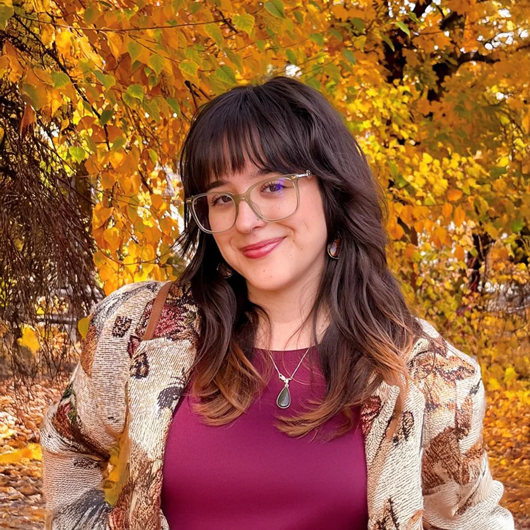 Aleena Mendoza smiling outdoors in front of autumn-colored trees, wearing glasses and a patterned cardigan.