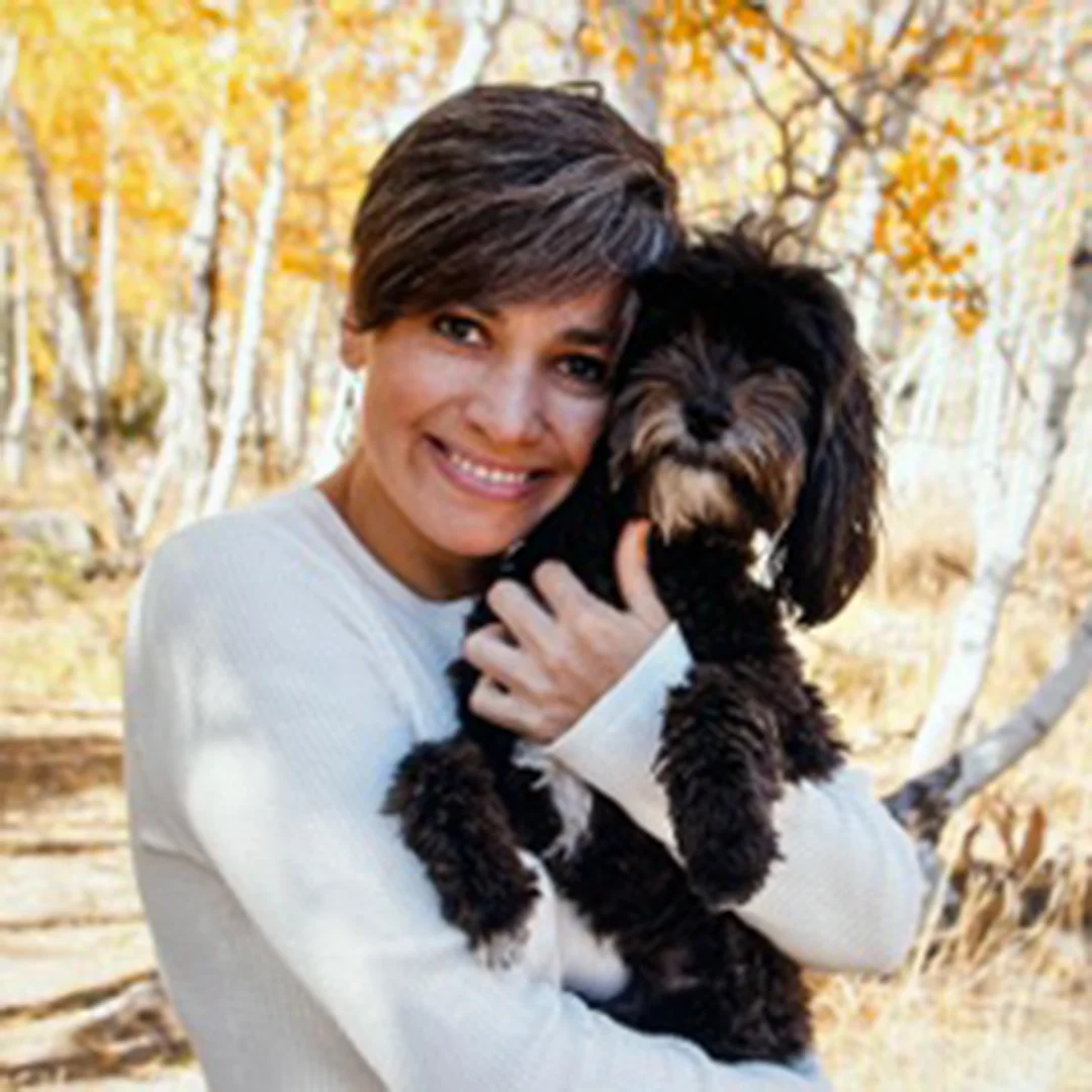 Beatriz Schaffert smiling outdoors in a wooded area, wearing a white top and holding a black dog.