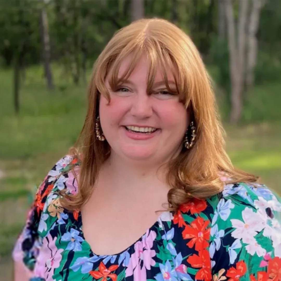 Brynne Paschal smiling outdoors in a colorful floral top with greenery in the background.