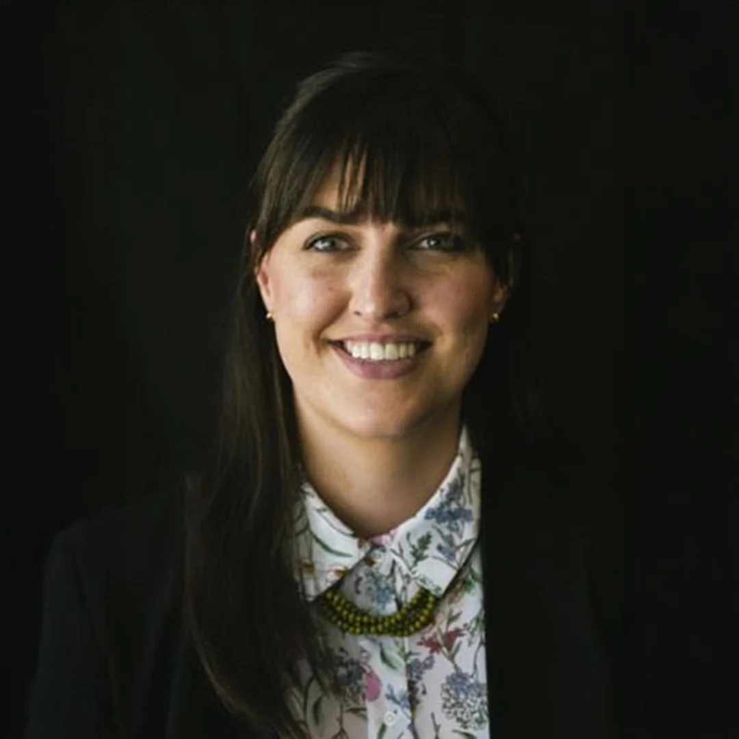 Camille Bailey smiling in front of a black background, wearing a floral blouse and dark blazer.