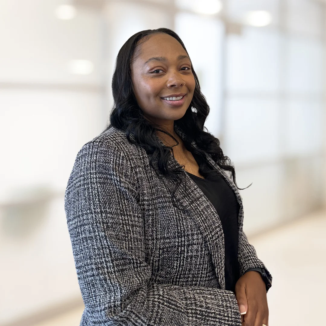 Janay Eustace smiling in a modern indoor space, wearing a textured gray blazer and black top.