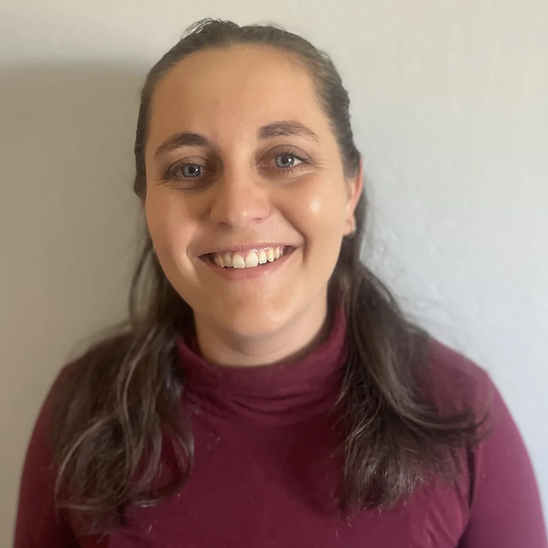 Kristen Gast smiling in front of a light-colored wall, wearing a maroon turtleneck top.