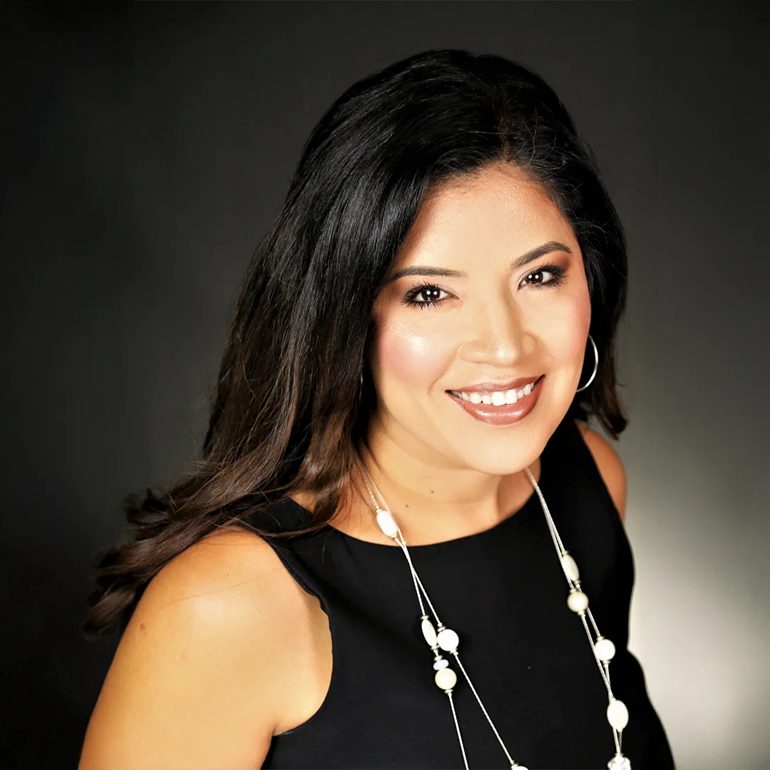 Lucy Hernandez smiling against a dark backdrop, wearing a black dress and pearl necklace.