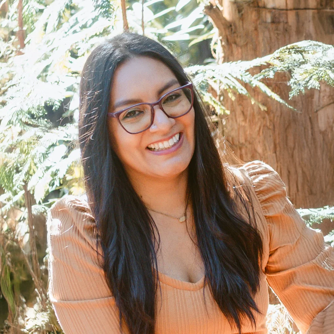 Maya Gutierrez smiling in a forested setting, wearing glasses and a rust-colored top.
