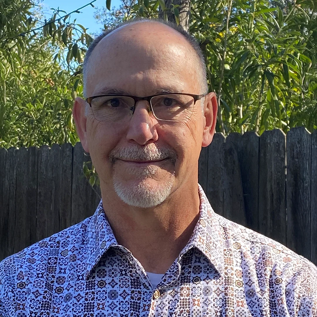 Michael Williams smiling outdoors in front of a wooden fence and trees, wearing glasses and a patterned button-up shirt.