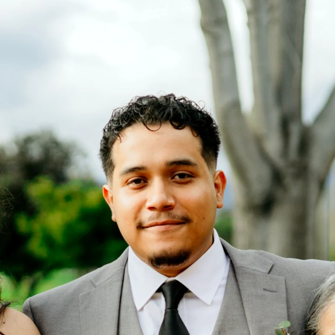 Nathanael Ruiz in a gray suit and black tie, smiling outdoors with trees in the background.