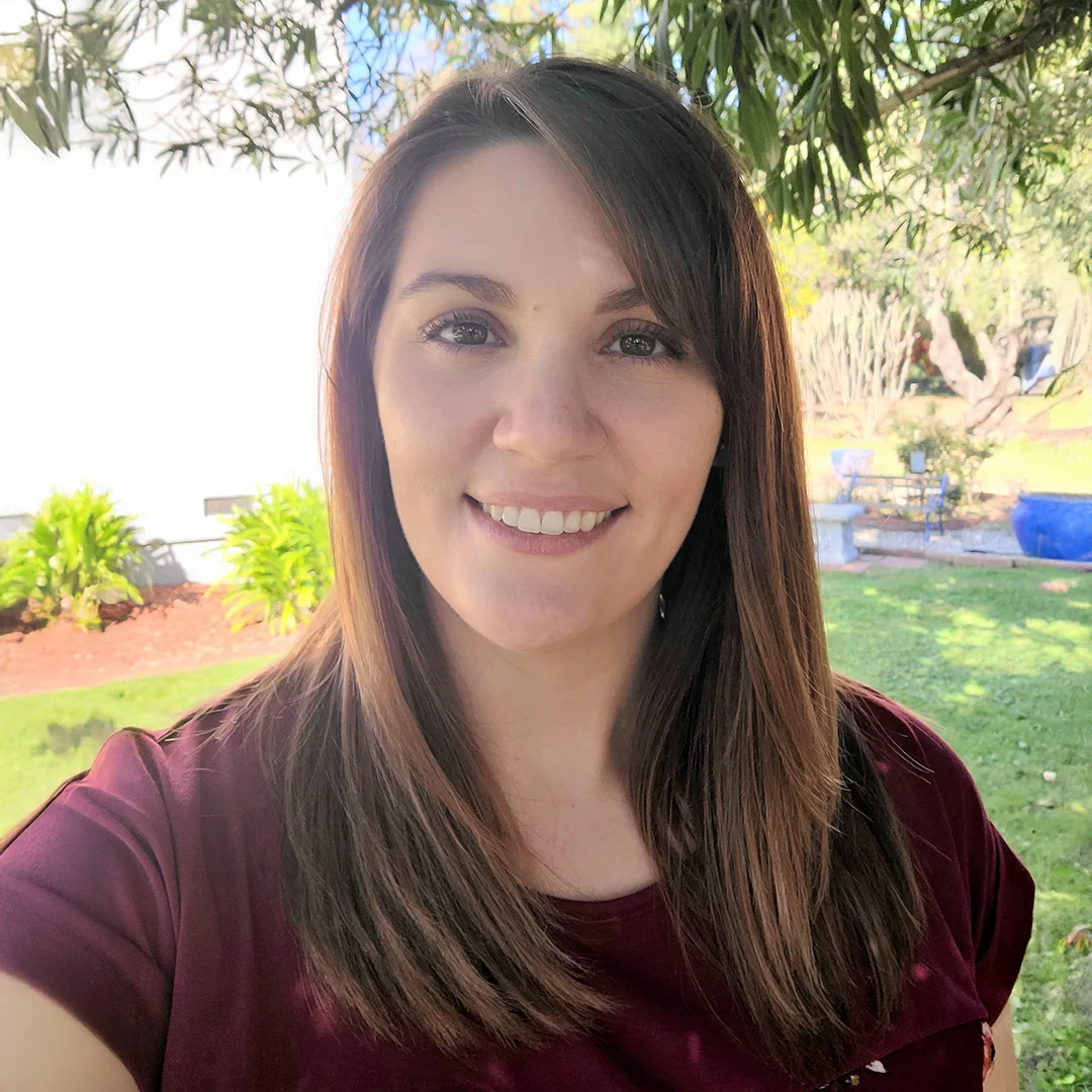 Regina Wong smiling outdoors in a sunny garden, wearing a maroon top with straight brown hair.