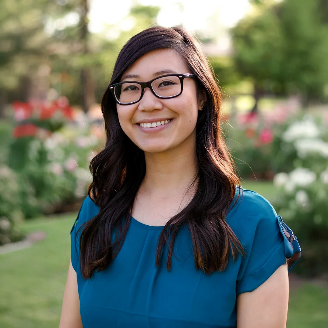 Serena Chau smiling outdoors in a garden, wearing glasses and a teal blouse.