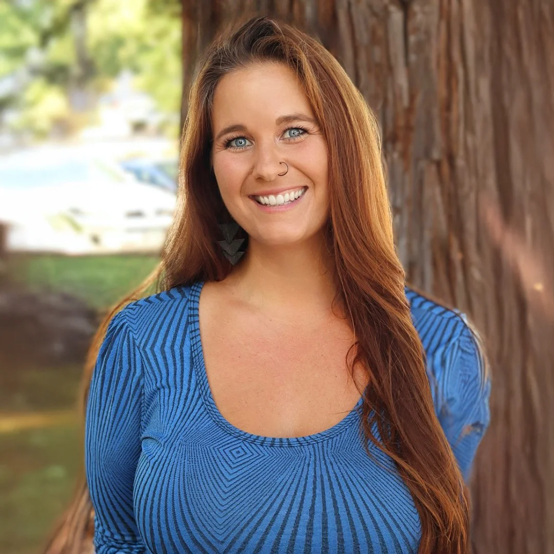 Tabitha Grier smiling in front of a redwood tree, wearing a blue patterned top with long red hair.