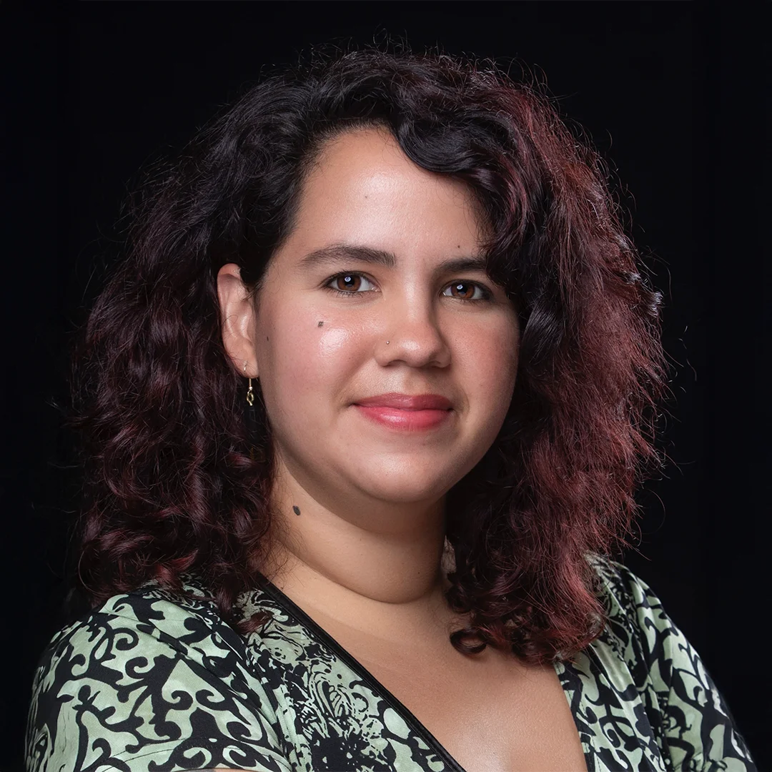 Victoria Hartman smiling in front of a black background, wearing a green and black patterned top with curly dark hair.