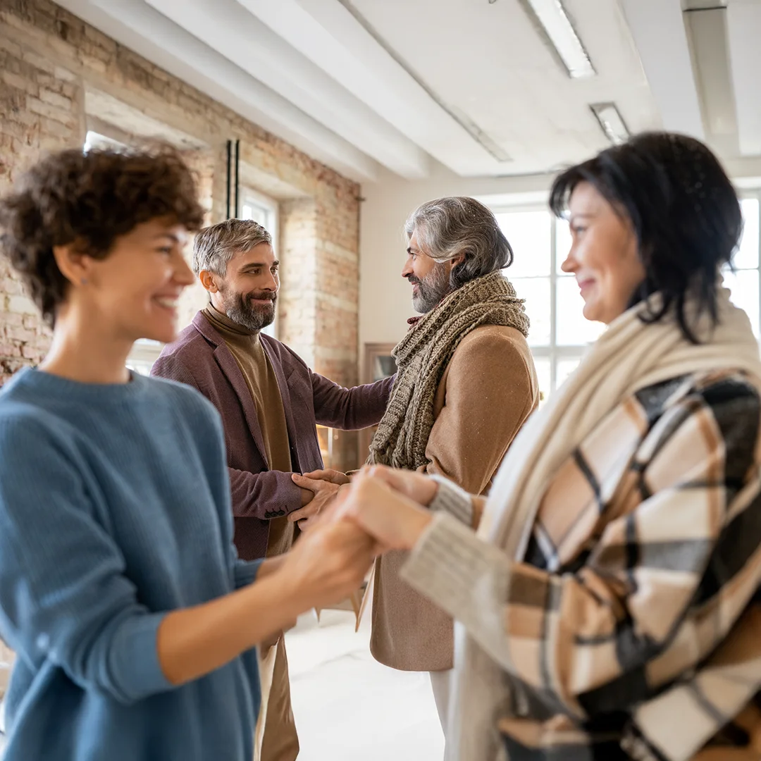 Two pairs of adults smiling and shaking hands in a cozy, sunlit room.