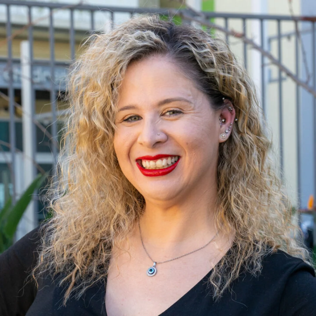 Laura Campana smiling outdoors in front of a building, wearing a black top and red lipstick with curly blonde hair.