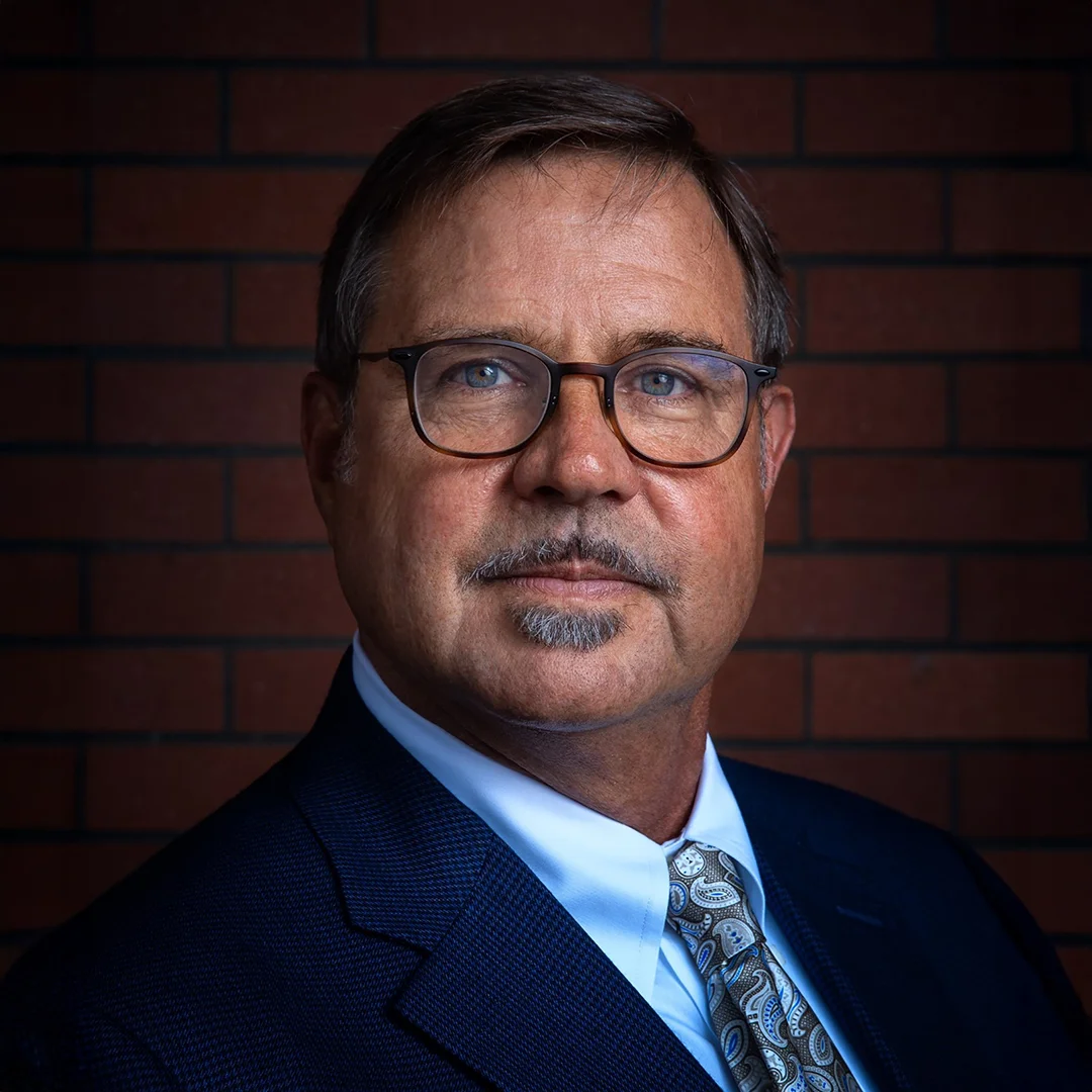 Ron Brown in a dark suit and patterned tie, posed in front of a brick wall with a serious expression.